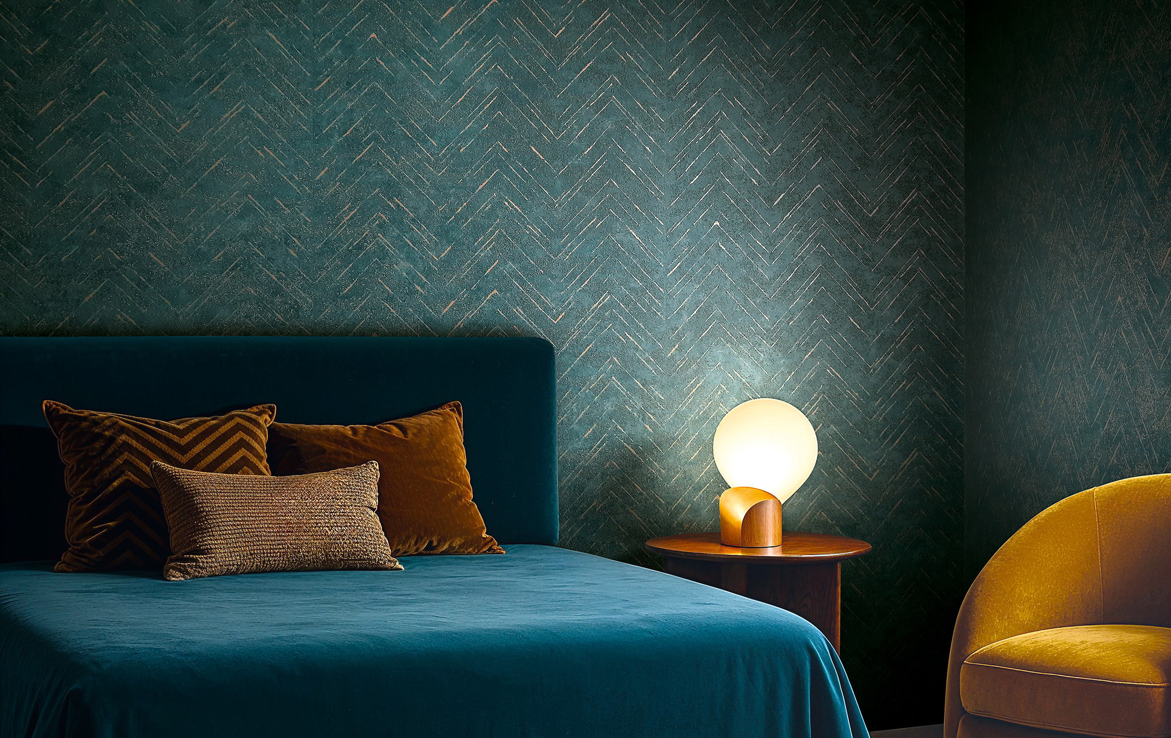 Schlafzimmer mit tiefgrüner Tapete in Fischgrätstruktur, Samtbett in Petrol, warmen Ockertönen und gedämpfter Lichtstimmung für ruhige Raumatmosphäre.